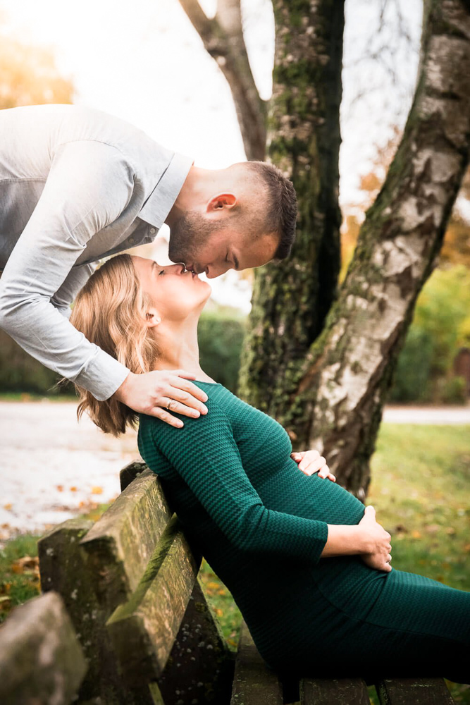 Die Schwangere sitzt auf einer Parkbank und bekommt einen Kuss von ihrem Mann, der hinter der Bank steht.