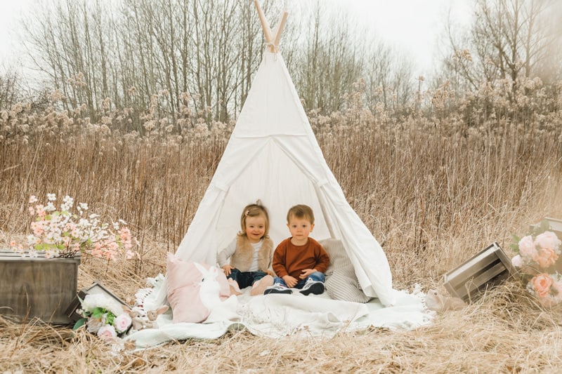 Zweijährige Zwillinge sitzen in einem Tipi-Zelt vor einem vertrocknetem Feld.
