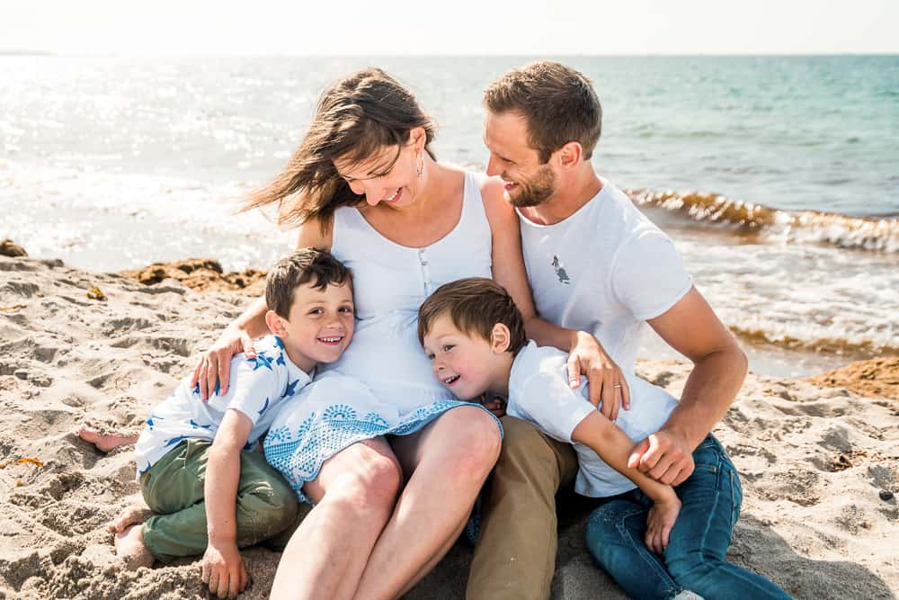 Familie sitzt am Strand und ihre zwei Söhne kuscheln den Babybauch.