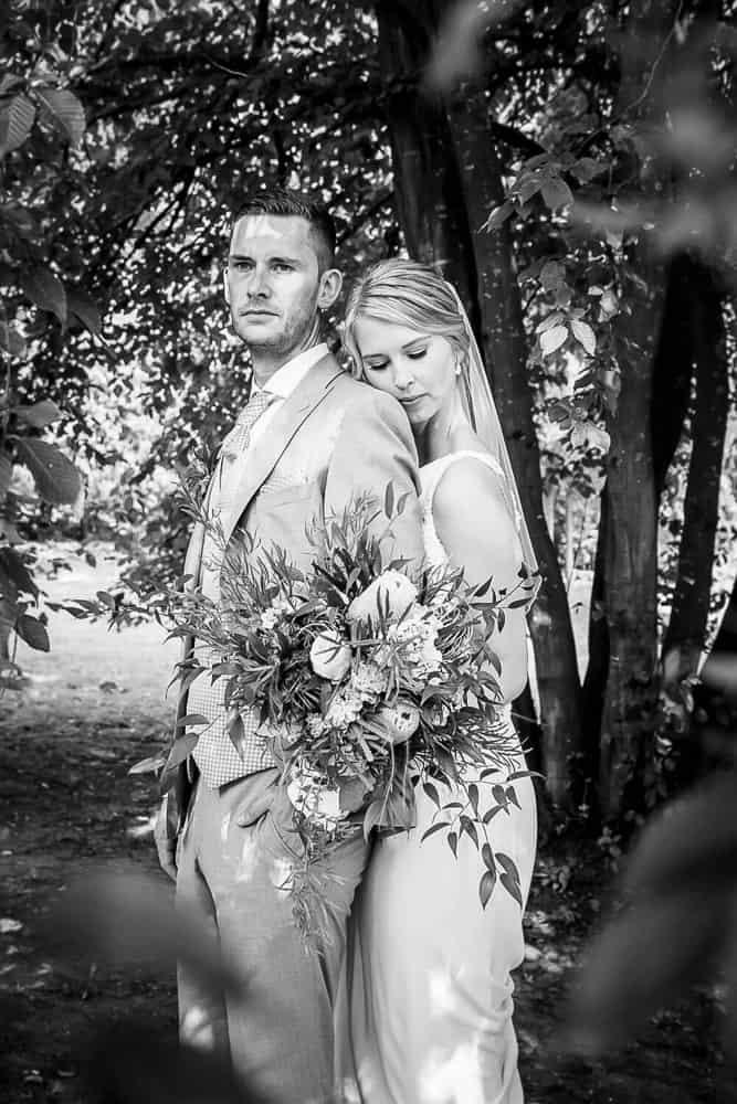 Schwarzweiß-Foto von Brautpaar unter einem Baum durch Blätter hindurch fotografiert. Die Braut steht am Rücken von ihrem Mann und kuschelt sich an. Hochzeitsfotograf Gut Gerdshagen bei Satow in Mecklenburg-Vorpommern.