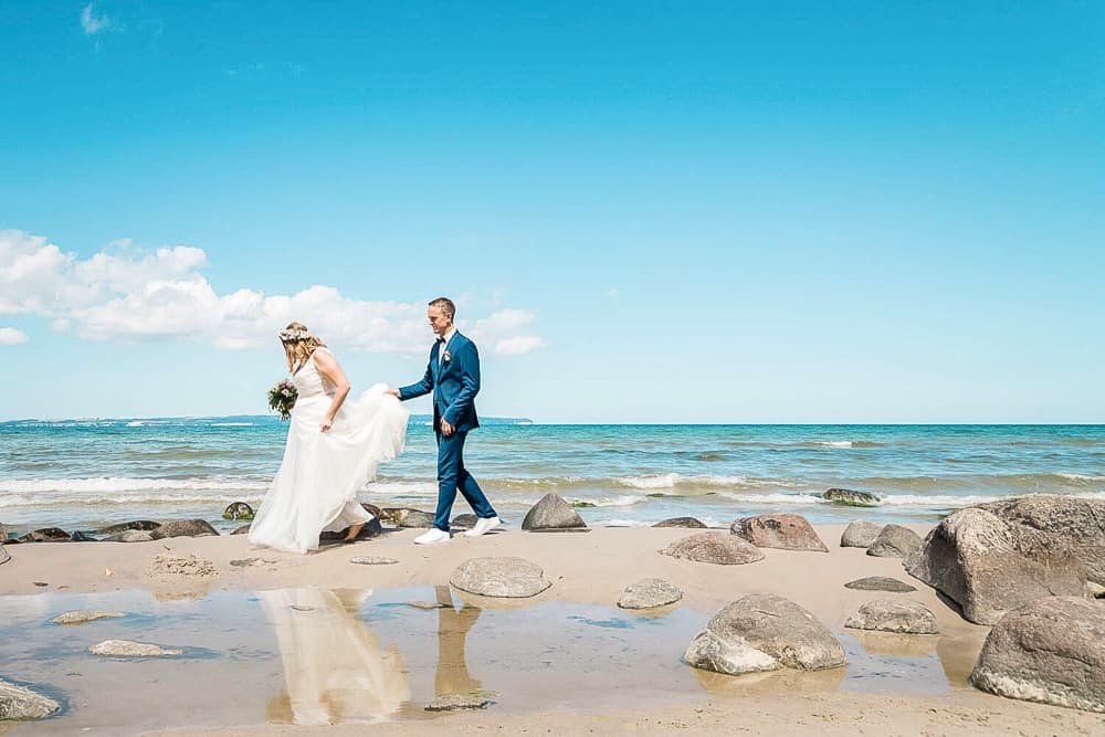 Brautpaar läuft am Strand von Binz entlang. Große Steine zieren die Kulisse bei strahlend blauem Himmel. Hochzeitsfotograf Rügen.
