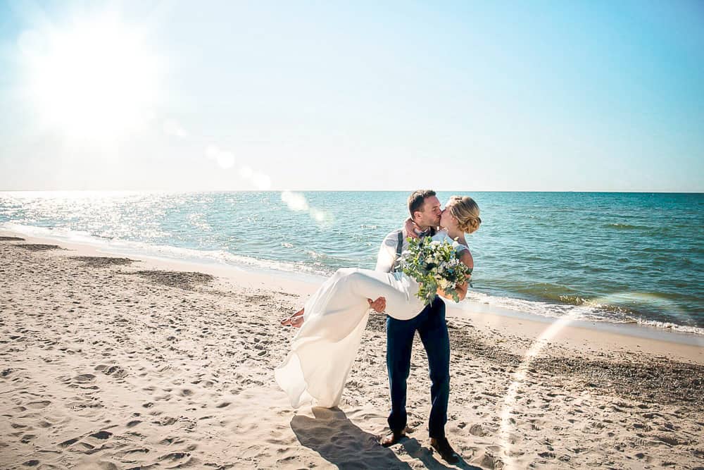 Der Bräutigam trägt seine Braut am Strand und küsst sie. Blauer Himmel, blaues Meer. Eine wunderschöne Kulisse für die Hochzeitsfotos mit Hochzeitsfotograf aus Dierhagen.