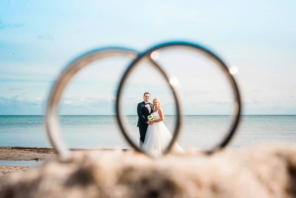 Ein kreatives Hochzeitsfoto: Die Eheringe im Vordergrund und das Brautpaar steht in diesen drinnen. Hochzeitsfoto am Strand von Warnemünde mit Hochzeitsfotograf aus Rostock.