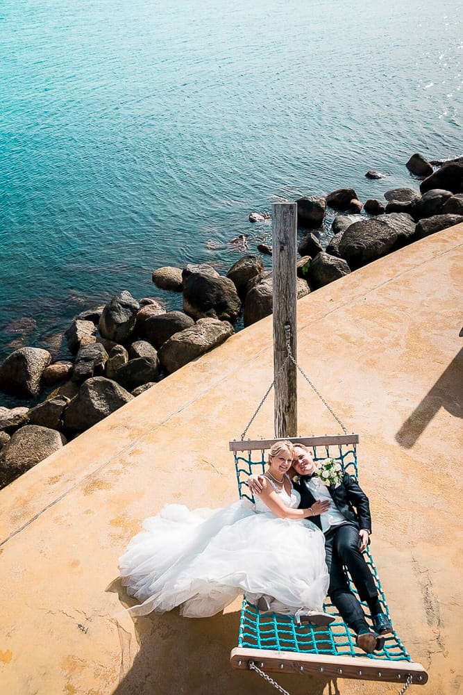 Brautpaar liegt in einer Hängematte und im Hintergrund ist die Ostsee zu sehen. Das Hochzeitsfoto wurde aus der Vogelperspektive aufgenommen. Hochzeitsfotograf Kellenhusen.