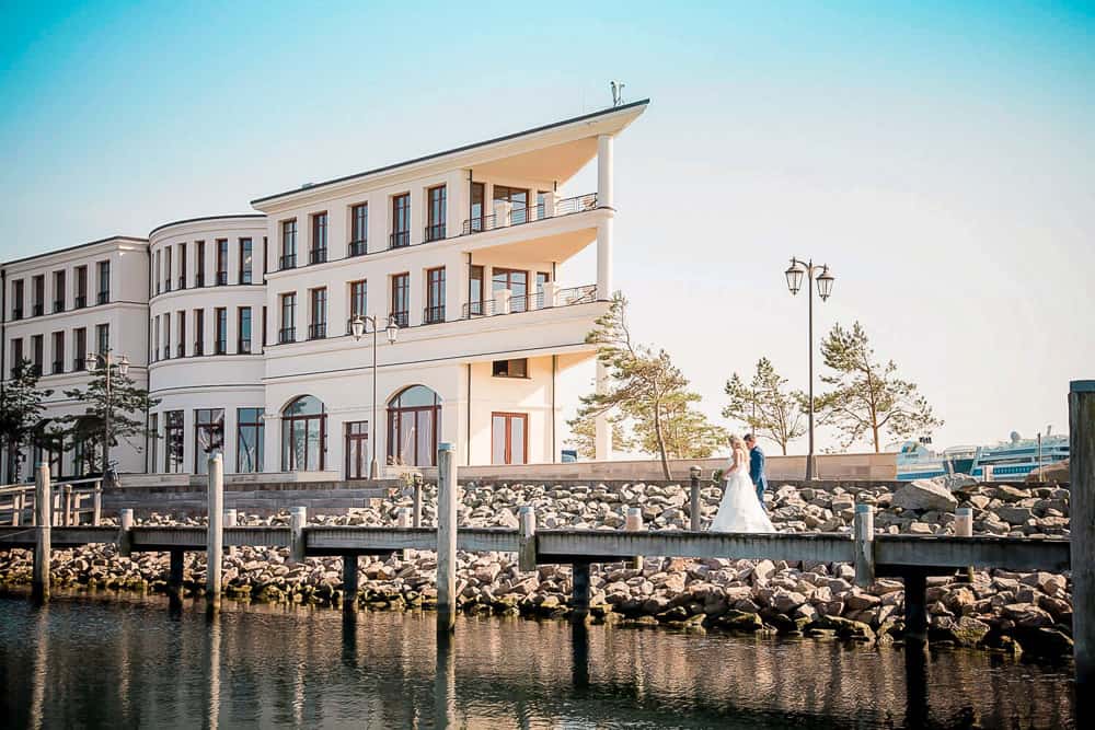 Hochzeitsfoto beim Brautpaar-Fotoshoong in Hohe Düne Markgrafenheide bei Warnemünde. Das Hochzeitspaar läuft auf einem Steg am Hafen entlang. Im Hintergrund sieht man das 5*-Hotel. Hochzeitsfotograf Rostock.