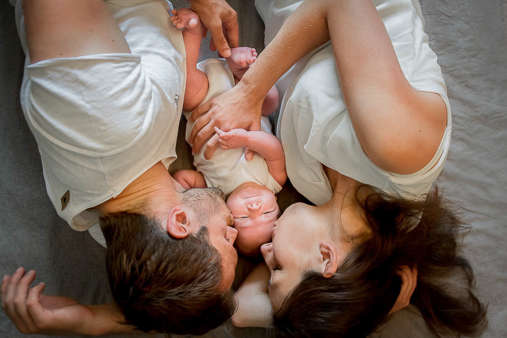 Eltern liegen auf dem Bett mit ihrem neugeborenem Baby zwischen ihnen und geben ihm ein Küsschen