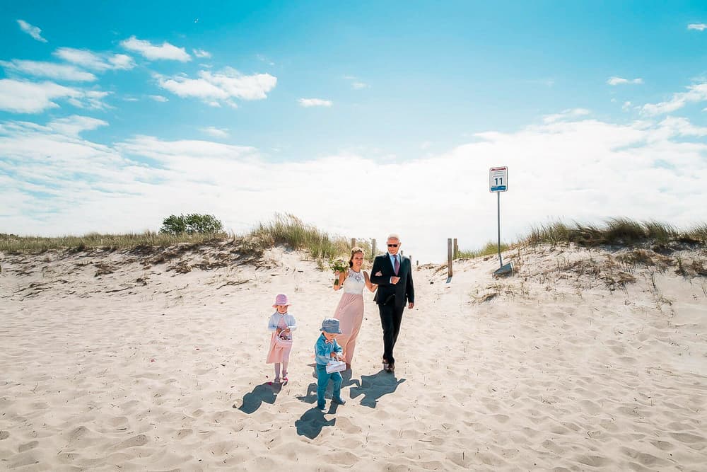 Braut, Brautpapa und die Blumenkinder laufen am Strand zur Trauung in Ahrenshoop am Strand. Heiraten an der Ostsee. Hochzeitsfotograf Darß.