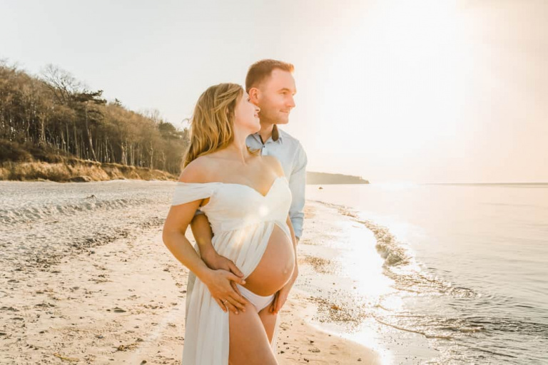 Schwangere Frau und ihr Mann stehen zusammen gekuschelt am Strand zur goldenen Stunde.