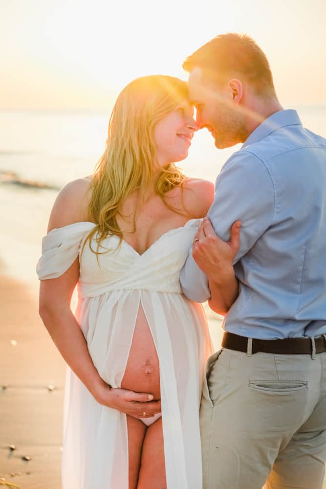 Schwangere Frau und ihr Mann schauen sich verliebt am Strand zum Sonnenuntergang an.