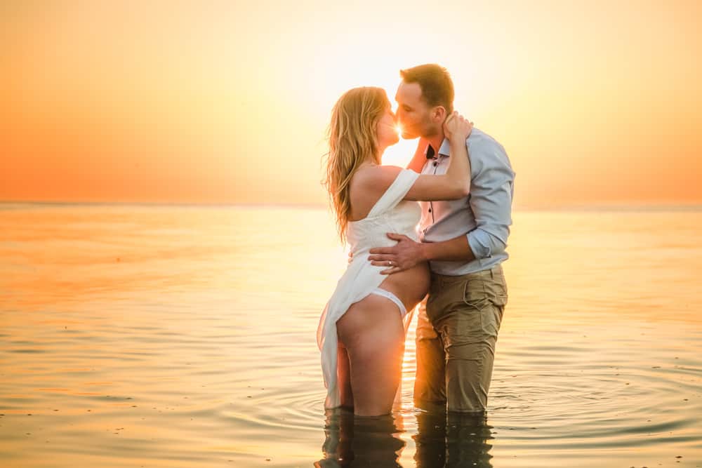 Schwangere Frau und Mann stehen im Wasser zum Sonnenuntergang und küssen sich.