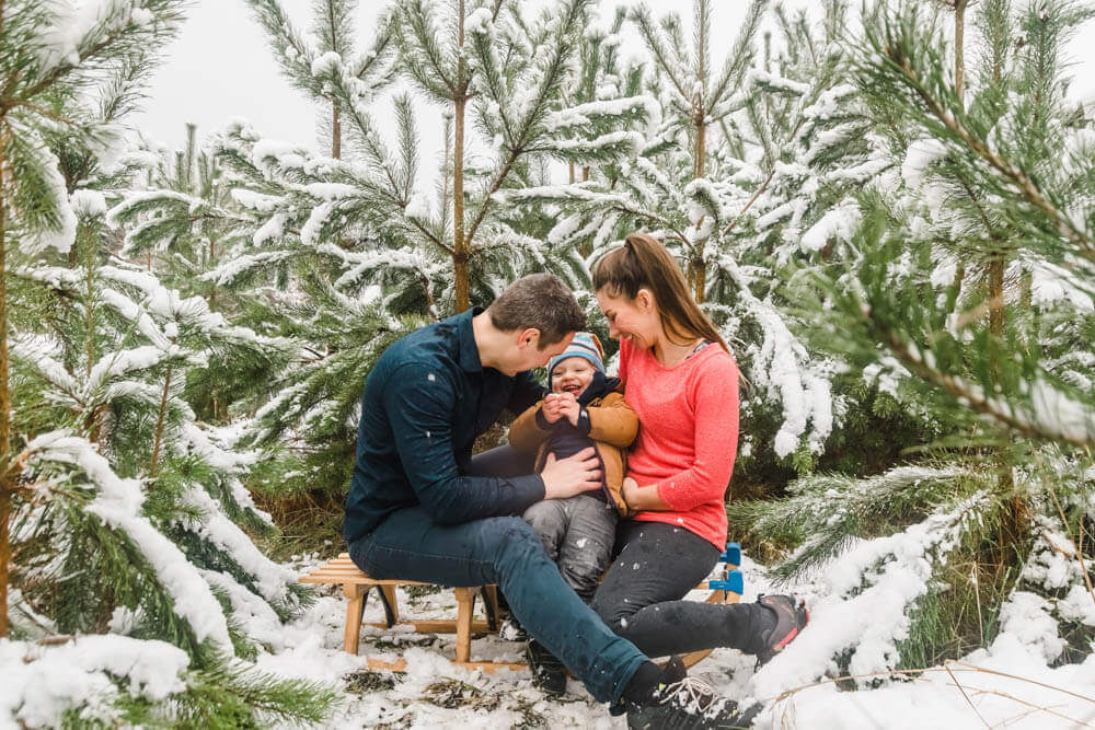 Mama und Papa mit Kleinkind auf einem Schlitten zwischen Tannenbäumen voller Schnee.