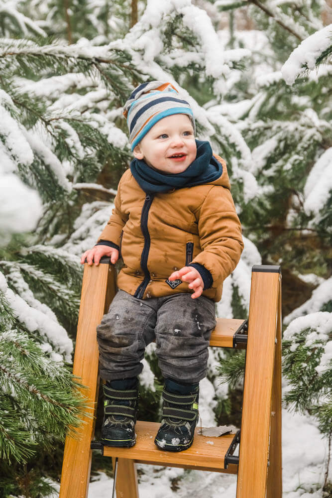 Kleinkind sitzt auf einer Leiter zwischen Tannen voller Schnee.