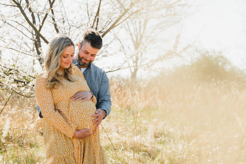 Schwangere Frau und ihr Mann sind stolz auf den Babybauch mit ihren Zwillingen. Sie trägt ein gelbes Kleid mit weißen Blumen und er trägt ein blaues legeres Hemd.