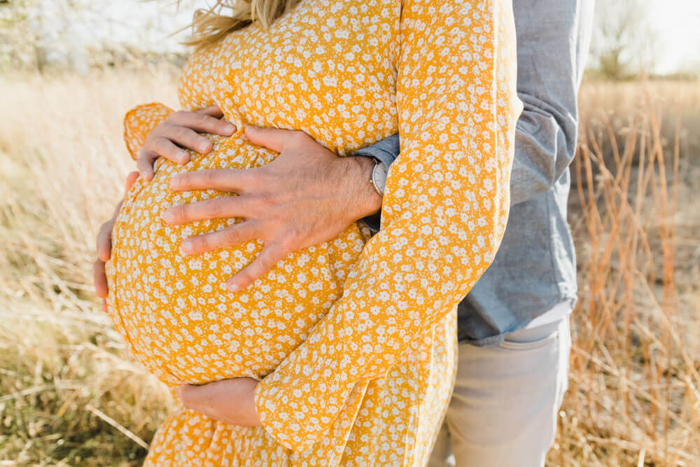 Aufnahme vom Babybauch mit Zwillingen in einem Feld. Der werdende Vater kuschelt den Bauch und die werdende Mama trägt ein gelbes Kleid mit weißen, kleinen Blumen.