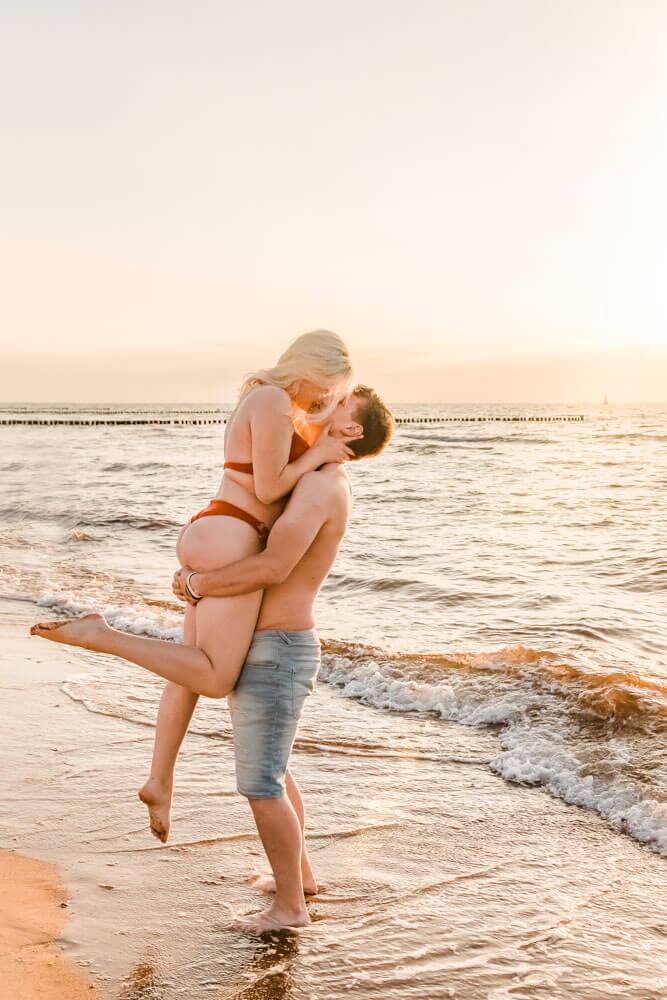 Freund hebt Freundin am Strand von Warnemünde zum Sonnenuntergang hoch, dabei küssen sie sich.