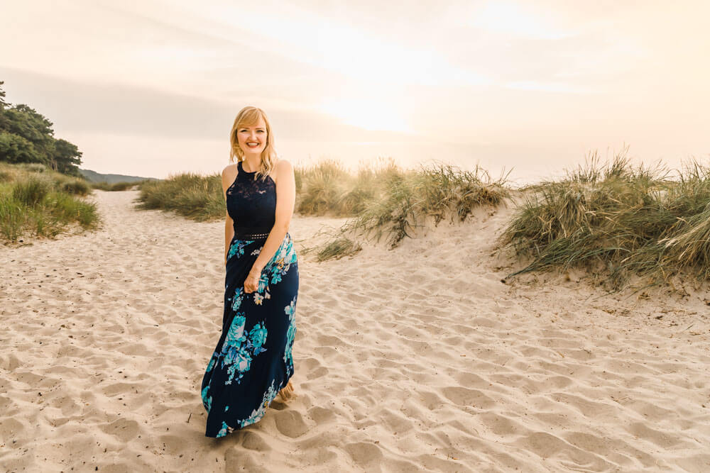 Blonde junge Frau trägt ein blaues Kleid mit Blumen und läuft am Strand von Warnemünde zwischen den Dünen entlang während der goldenen Stunde