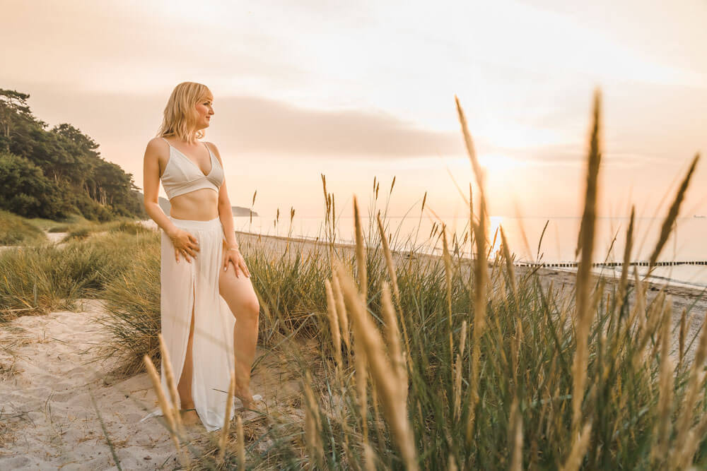 Blonde junge Frau steht in den Dünen von Warnemünde und schaut zum Sonnenuntergang auf die Ostsee