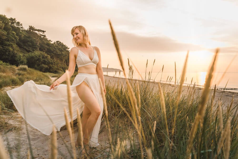 Junge blonde Frau zur goldenen Stunde am Strand von Warnemünde zwischen den Dünen mit weißem Outfit