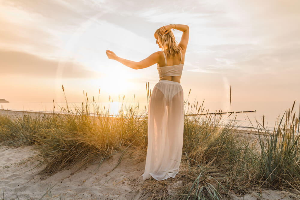 Junge blonde Frau mit weißem Outfit steht auf den Dünen beim Strand von Warnemünde zum Sonnenuntergang
