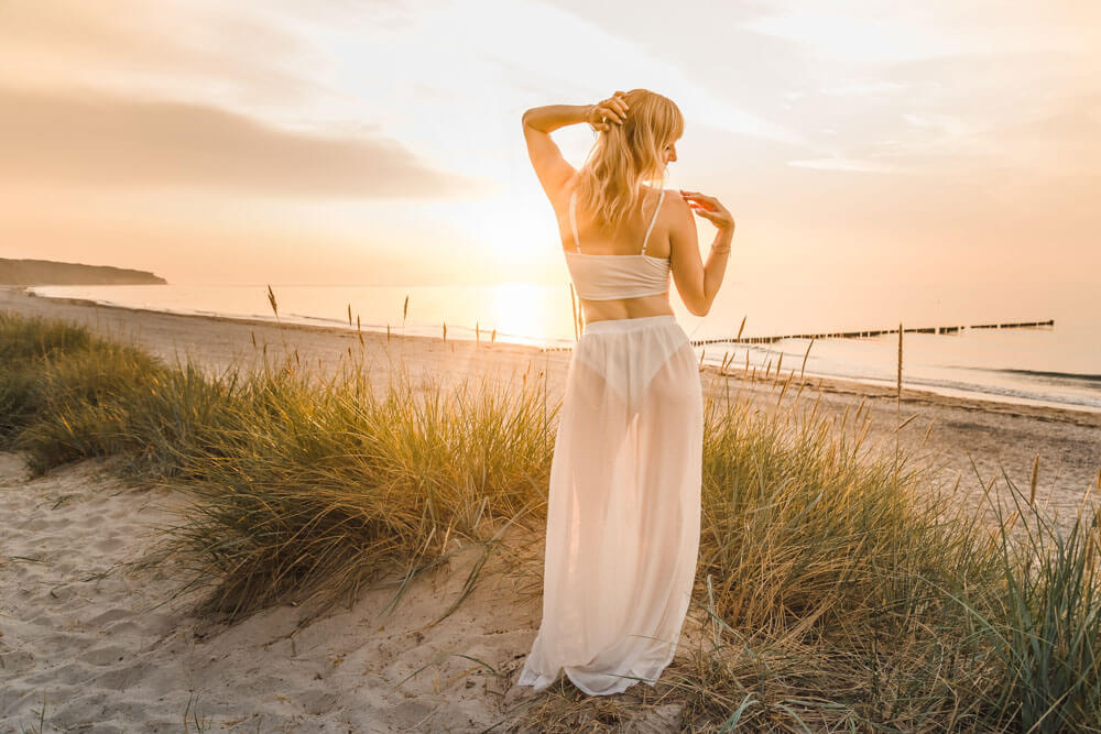 Blonde junge Frau mit weißem Rock in den Dünen am Strand von Warnemünde zur goldenen Stunde