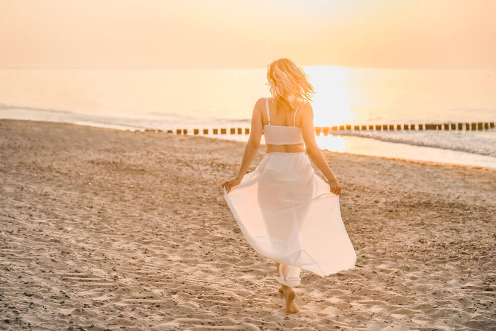 Junge blonde Frau mit weißem Outfit rennt am Strand von Warnemünde zur Sonne