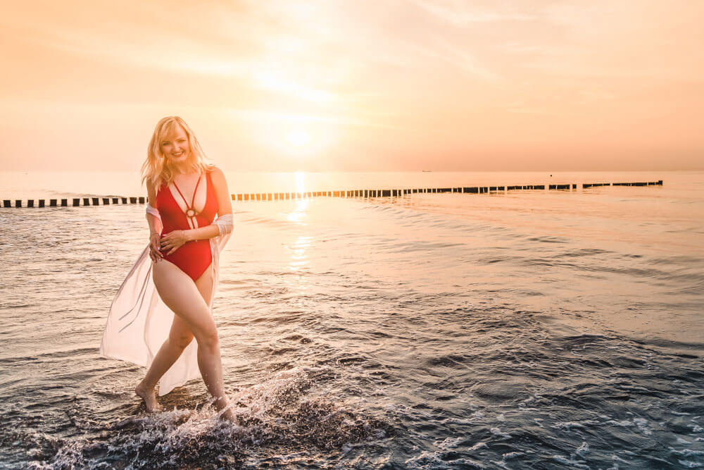Junge Frau rennt im roten Bikini durch das Wasser der Ostsee beim Sonnenuntergang in Warnemünde.
