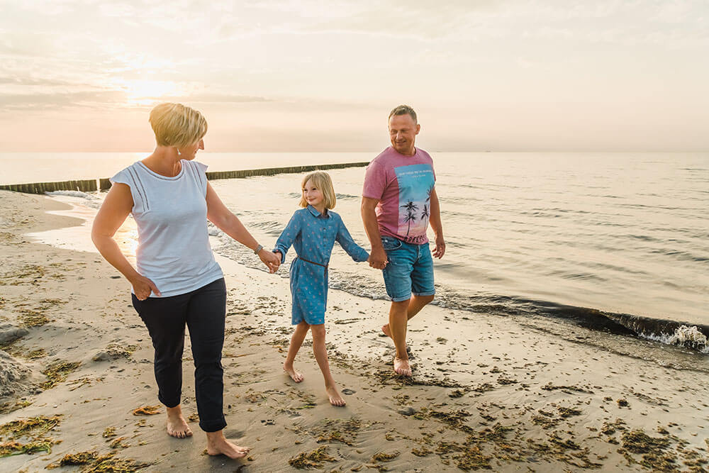 Familie macht einen Spaziergang an der Ostsee zum Sonnenuntergang. Die Eltern halten ihre Tochter zwischen sich an den Händen.