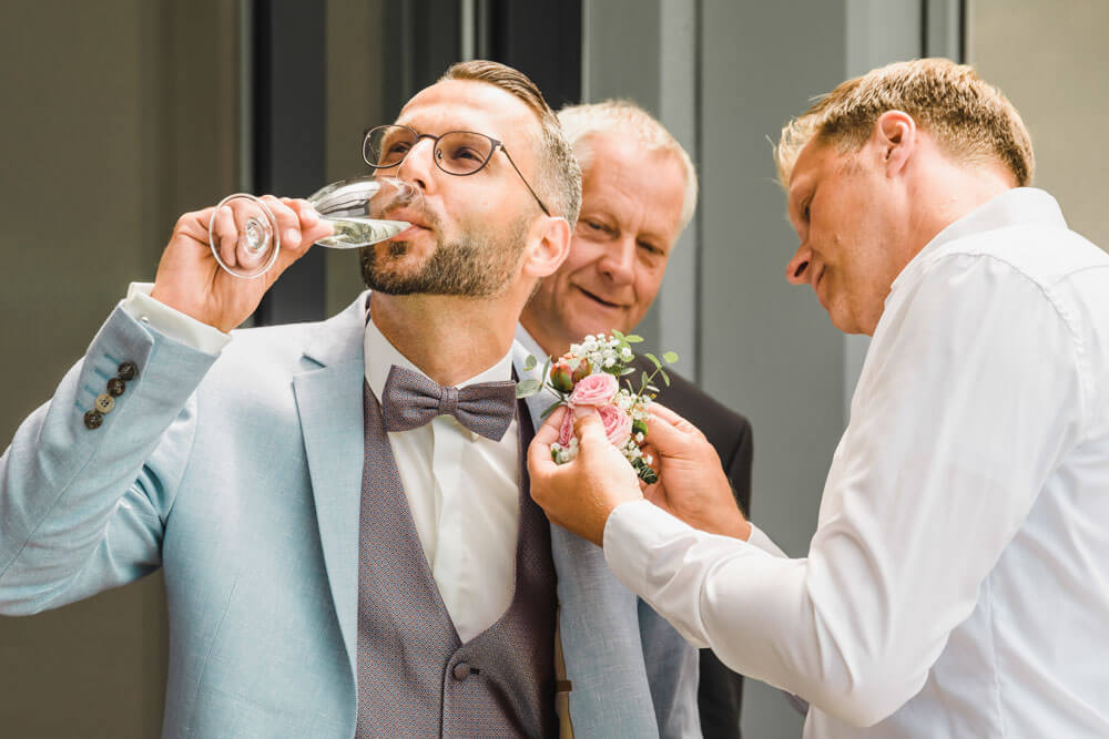 Getting Ready vom Bräutigam in einem Ferienhaus in Zingst auf dem Darss an der Ostsee. Der Bruder bringt die Ansteckblume beim Bräutigam an, während dieser einen Sekt trinkt.