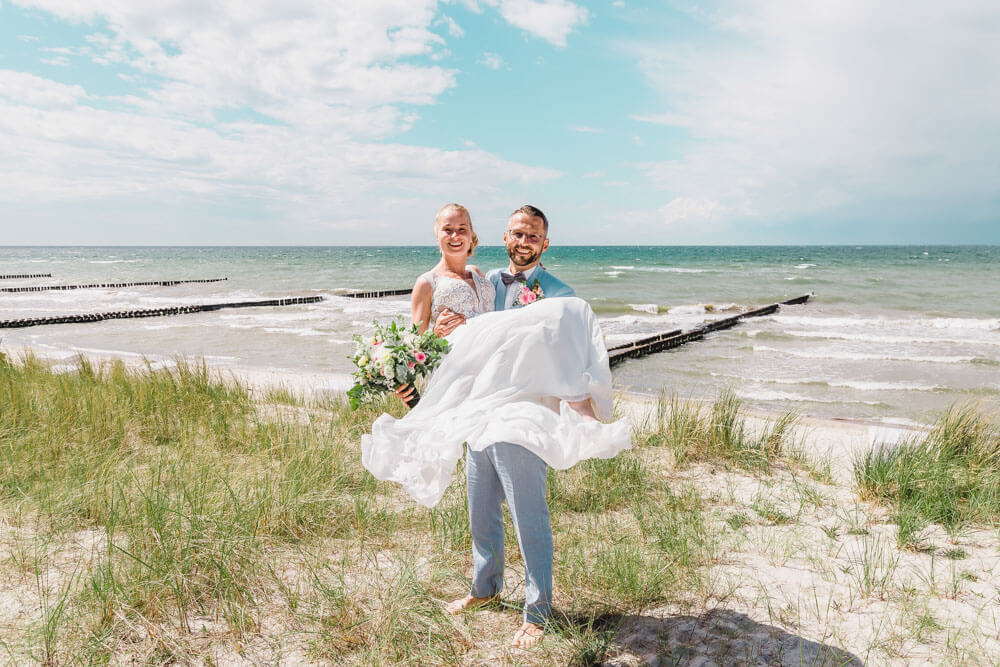 Bräutigam trägt Braut in den Dünen mit wunderschöner Kulisse vom Strand. Brautpaarfotos am Ostseestrand auf dem Darss in Mecklenburg-Vorpommern. Hochzeitsfotograf an der Ostsee (Markgrafenheide, Graal-Müritz, Dierhagen, Ahrenshoop, Wustrow, Prerow, Zingst).