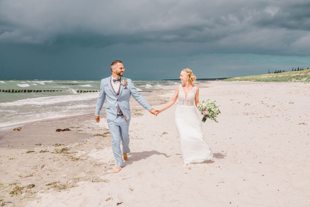 Brautpaar läuft am Strand entlang. Hinter ihnen ist ein Unwetter. Brautpaarfotos am Ostseestrand auf dem Darss in Mecklenburg-Vorpommern. Hochzeitsfotograf an der Ostsee (Markgrafenheide, Graal-Müritz, Dierhagen, Ahrenshoop, Wustrow, Prerow, Zingst).