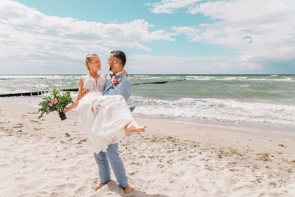 Bräutigam trägt seine Braut am Strand. Brautpaarfotos am Ostseestrand auf dem Darss in Mecklenburg-Vorpommern. Hochzeitsfotograf an der Ostsee (Markgrafenheide, Graal-Müritz, Dierhagen, Ahrenshoop, Wustrow, Prerow, Zingst).
