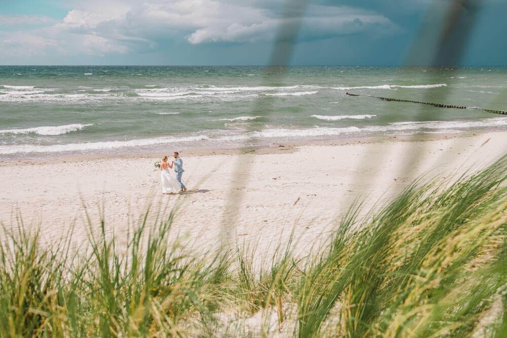 Das Hochzeitspaar am menschenleeren Strand aus den Dünen fotografiert. Brautpaarfotos am Ostseestrand auf dem Darss in Mecklenburg-Vorpommern. Hochzeitsfotograf an der Ostsee (Markgrafenheide, Graal-Müritz, Dierhagen, Ahrenshoop, Wustrow, Prerow, Zingst).