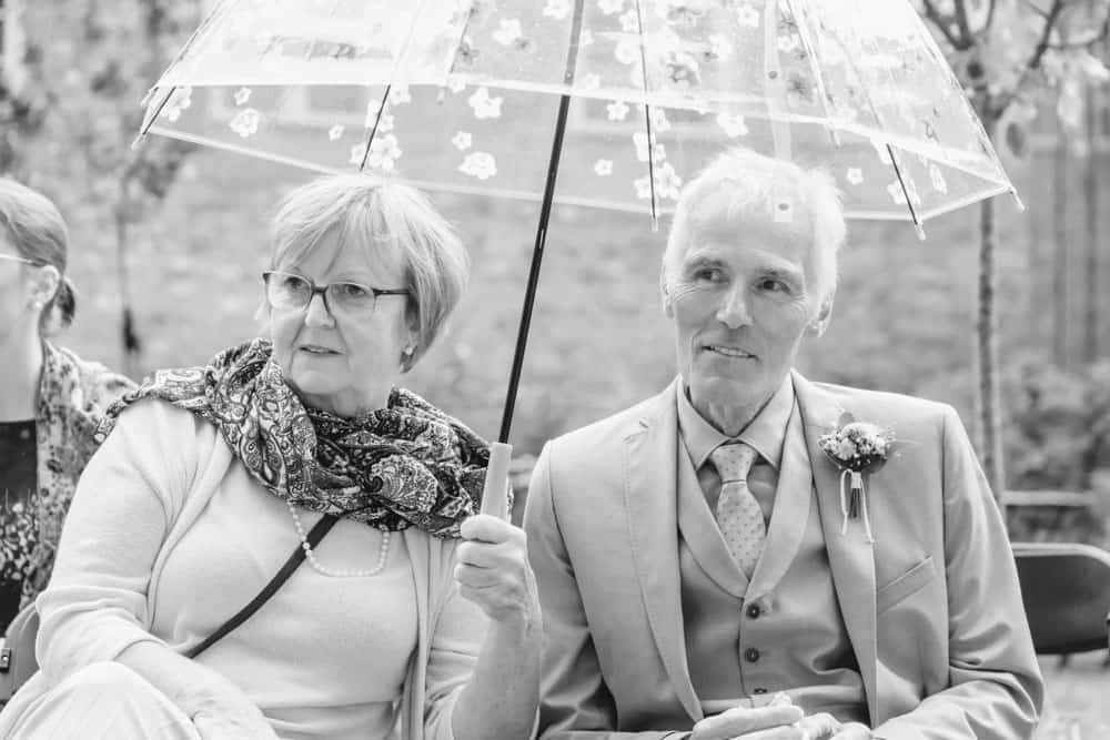 Schwarzweißfoto. Die Eltern vom Brautpaar sitzen unter einem durchsichtigen Schirm während der Hochzeit in Ribnitz-Damgarten.