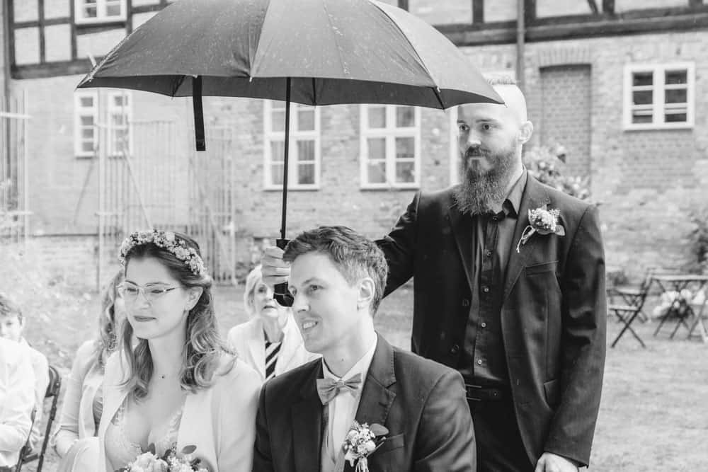 Schwarzweißfoto. Der Trauzeuge hält dem Hochzeitspaar einen Schirm, damit sie während der Trauung nicht nass werden.