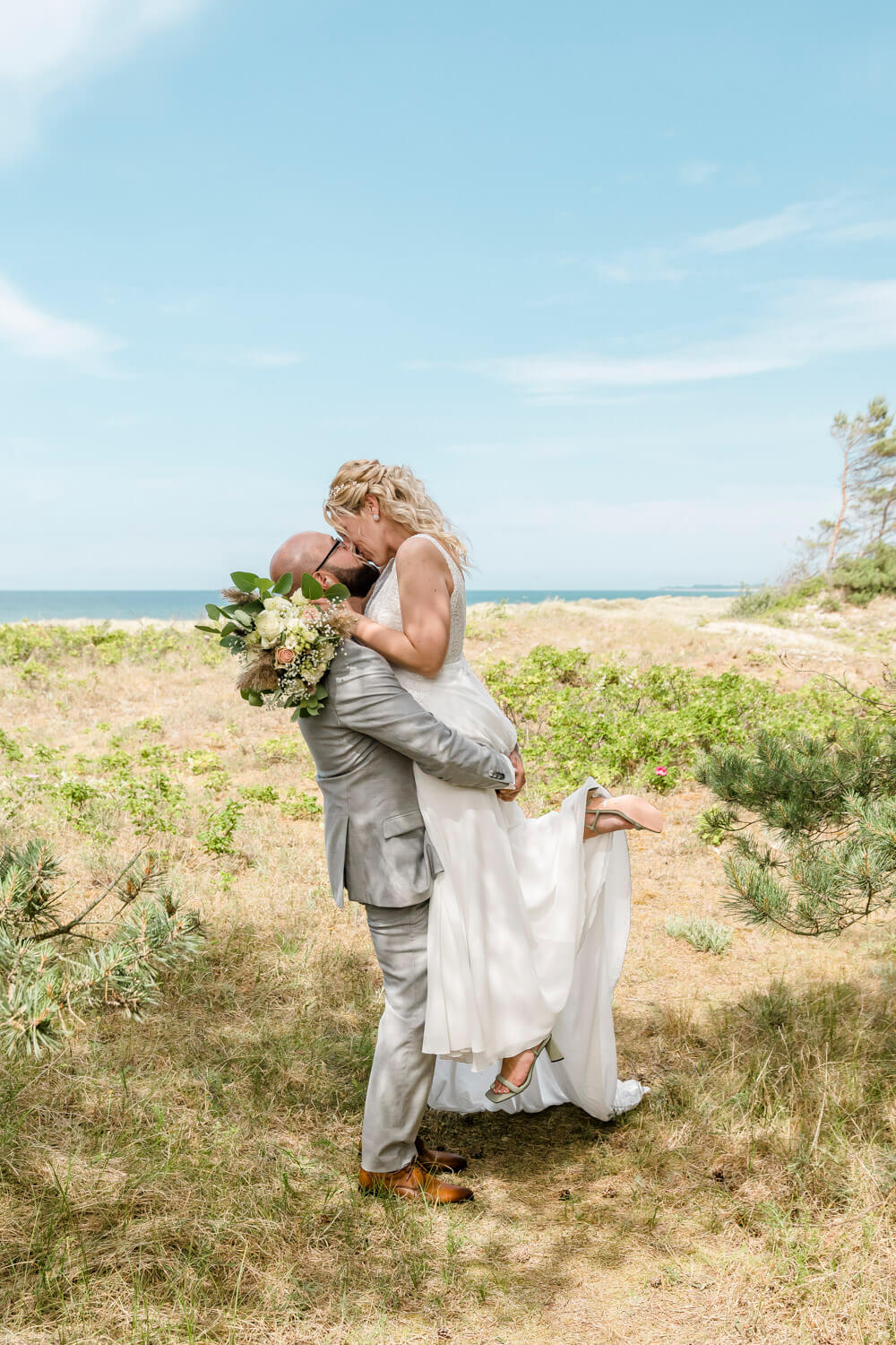 Der Ehemann hebt seine Braut hoch und küsst sie. Hochzeitsfoto mit Ostsee-Kulisse.