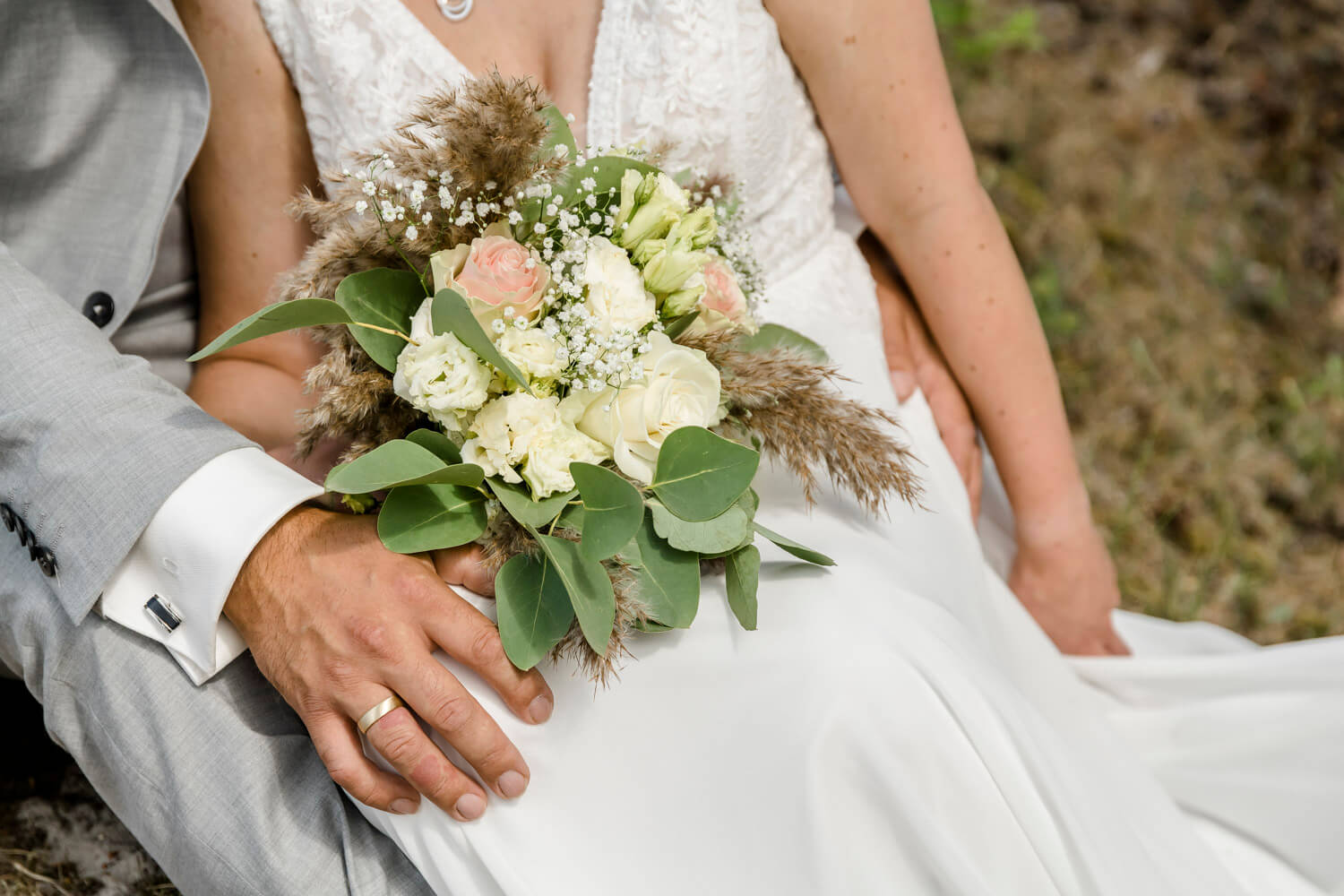 Detailfoto von der Hand des Bräutigams und dem modernen Brautstrauß mit Pampasgras und Rosen.