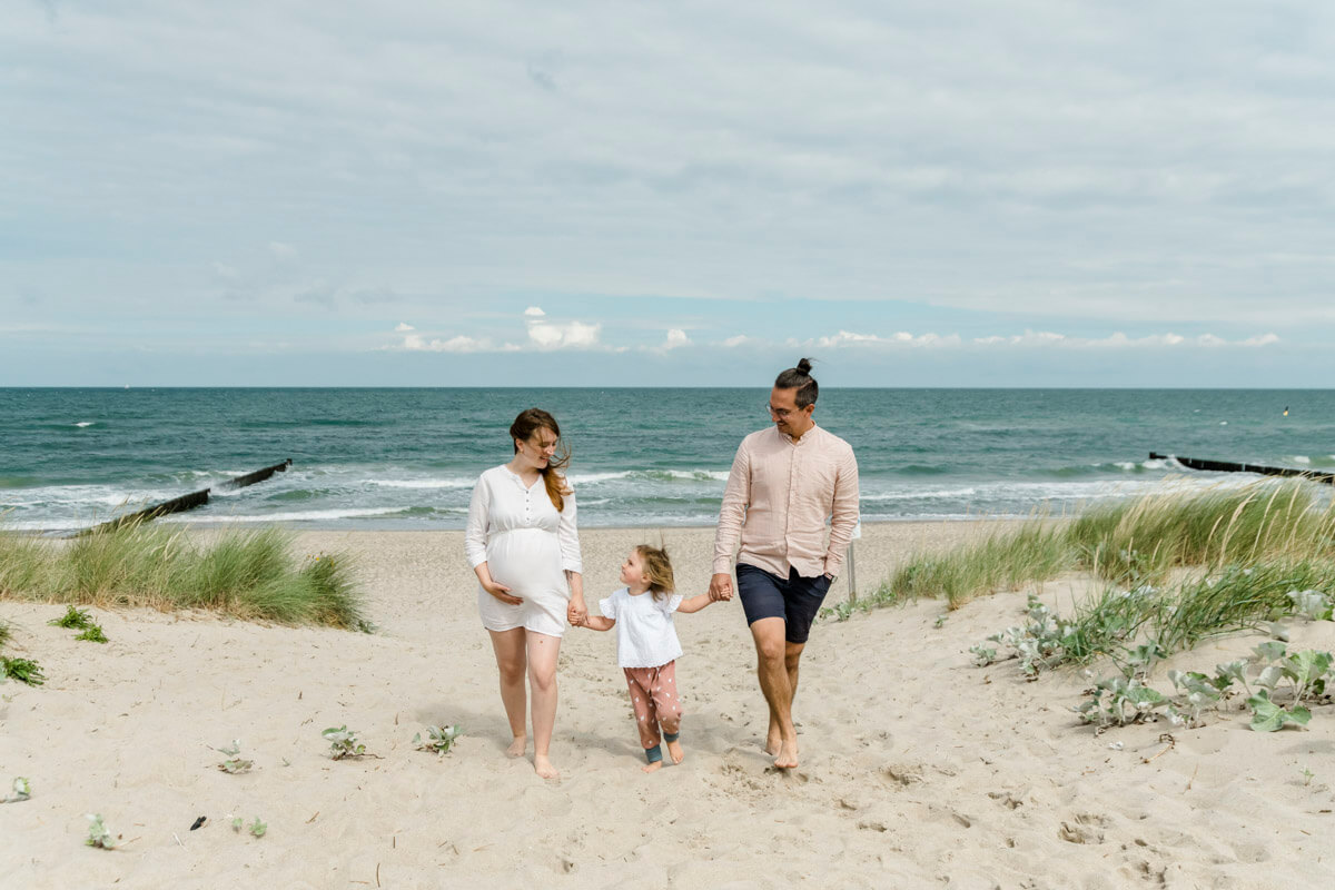 Babybauch-Fotoshooting in Graal-Müritz am Strand. Mama, Papa und Tochter laufen den Strandaufgang hoch - im Hintergrund ist die Ostsee zu sehen.