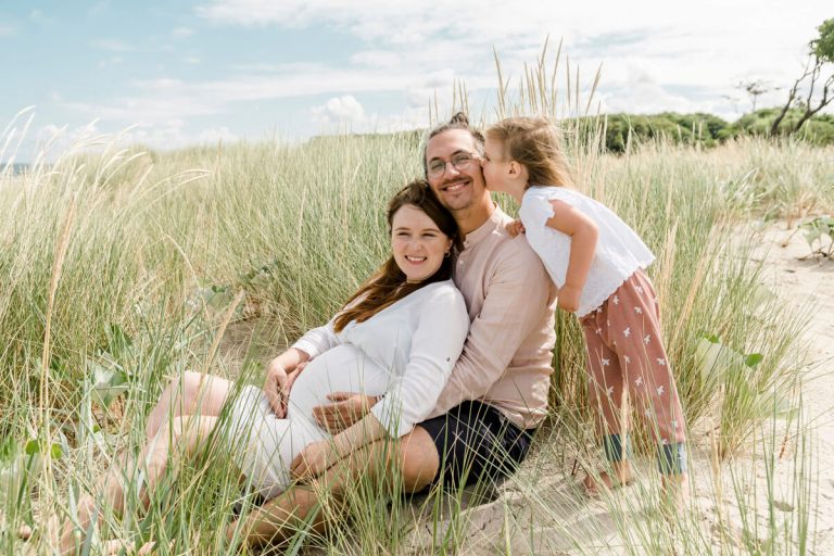 Babybauchfotos am Strand von Graal-Müritz mit Fotograf aus Ribnitz-Damgarten. Die Eltern sitzen in den Dünen und die große Schwester gibt dem Papa einen Kuss auf die Wange.