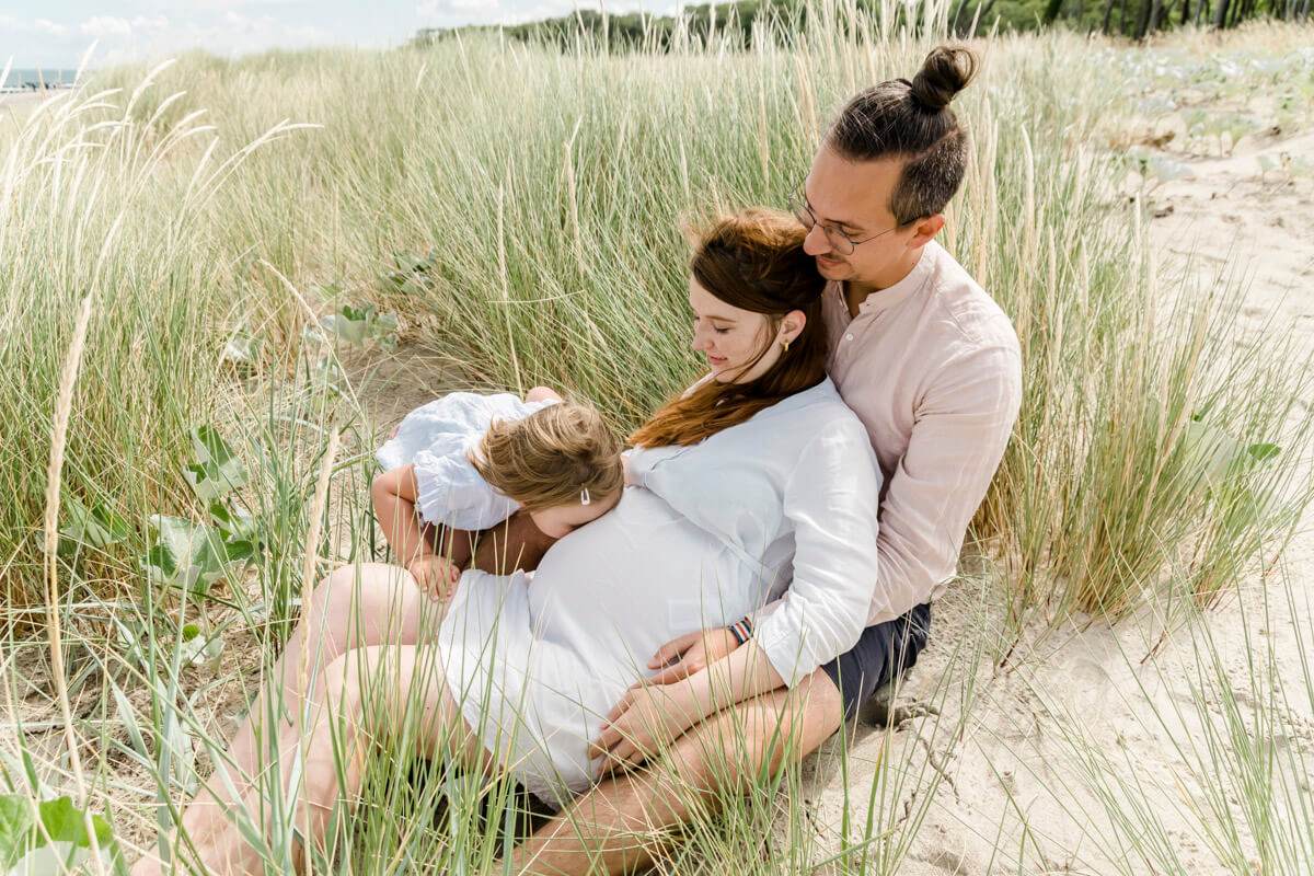 Schwangerschaftsfotos am Ostseestrand von Graal-Müritz. Mama und Papa sitzen in den Dünen. Die große Schwester gibt dem Babybauch einen Kuss.