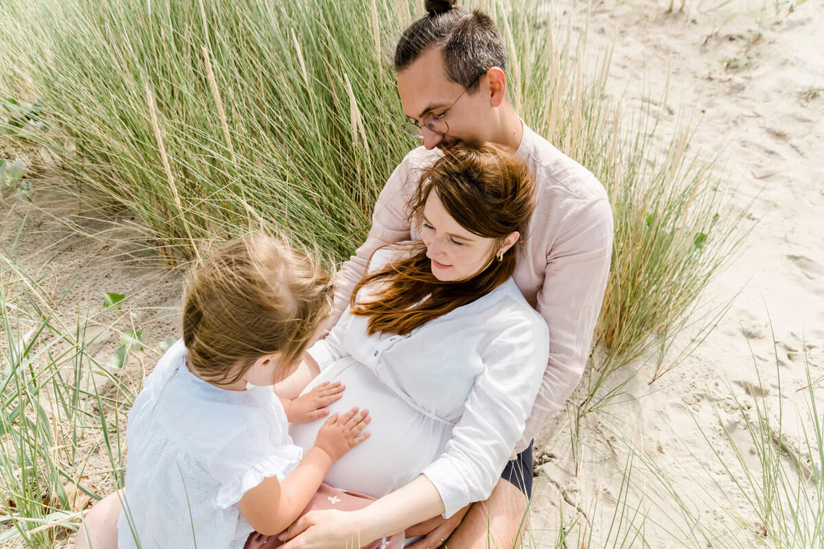 Vorfreude auf den Nachwuchs. Familie sitzt in den Dünen am Strand und genießen den Moment mit Babybauch.