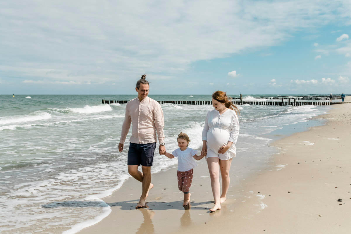 Familie läuft am Strand entlang. Sie halten ihre Tochter an den Händen. Die Mama ist schwanger. Fotograf Graal-Müritz.