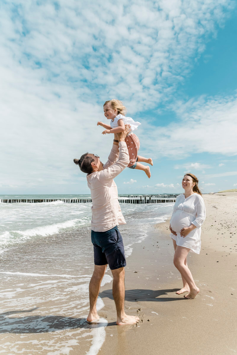 Familienfotos am Strand von Graal-Müritz. Der Papa wirft die Tochter in die Luft. Die Mama mit Babybauch steht daneben und freut sich.