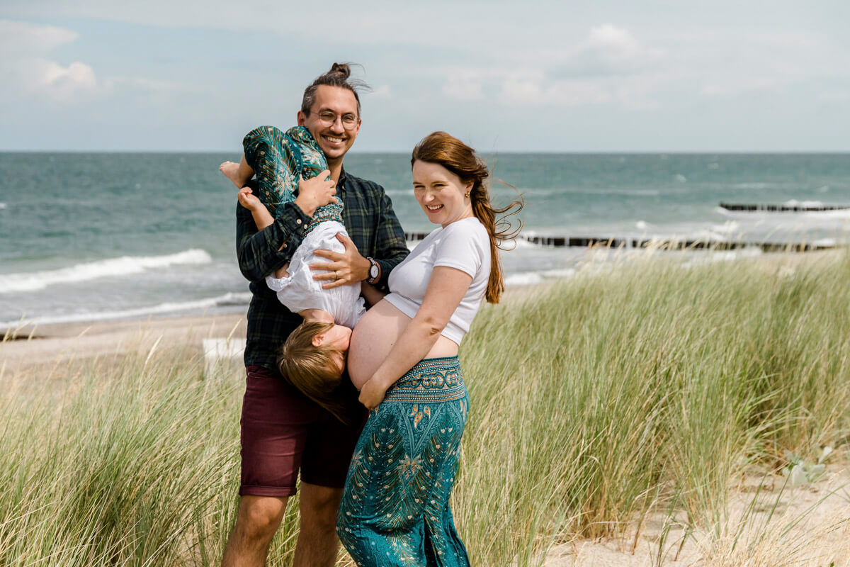 Der Papa hebt die Tochter verkehrt herum hoch, welche dem Babybauch von der Mama einen Kuss gibt. Lebendige und lustige Familienfotos am Strand von GRaal-Müritz.