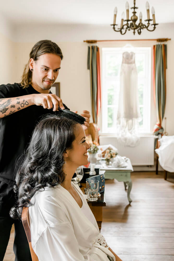 Der Friseur stylt die Haare für die Braut während dem Getting Ready im Gut Klein Nienhagen in der Nähe von Rostock. Im Hintergrund hängt das Brautkleid.