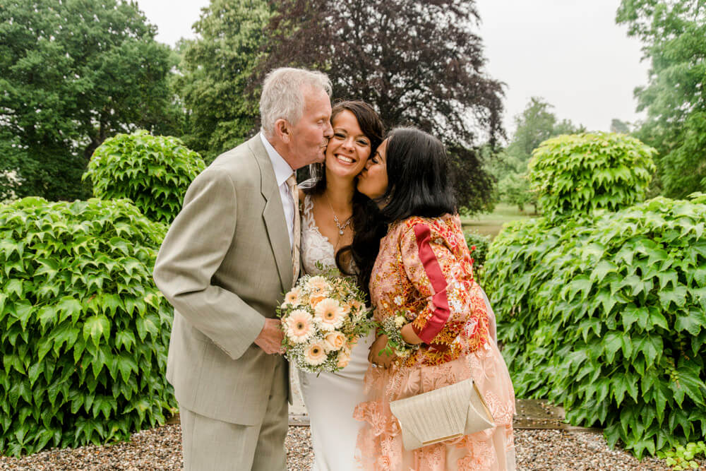 Gruppenfoto zur Hochzeit: Eltern der Braut geben ihr ein Küsschen auf die Wange