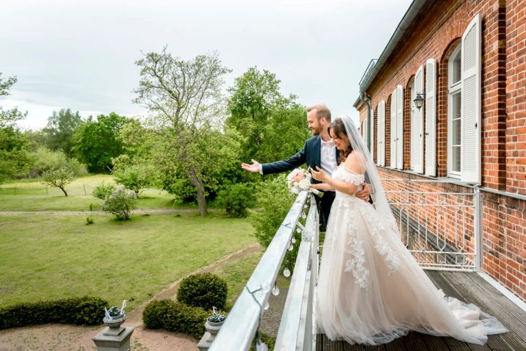 Das Brautpaar steht zur Hochzeit auf der Terrasse vom Gut Pütnitz in Ribnitz-Damgarten und winkt ihren Hochzeitsgästen im Garten zu. Hochzeitsfotograf aus der Region.