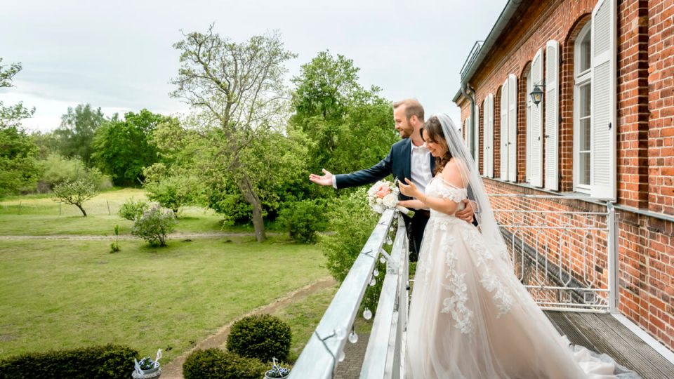 Das Brautpaar steht zur Hochzeit auf der Terrasse vom Gut Pütnitz in Ribnitz-Damgarten und winkt ihren Hochzeitsgästen im Garten zu. Hochzeitsfotograf aus der Region.