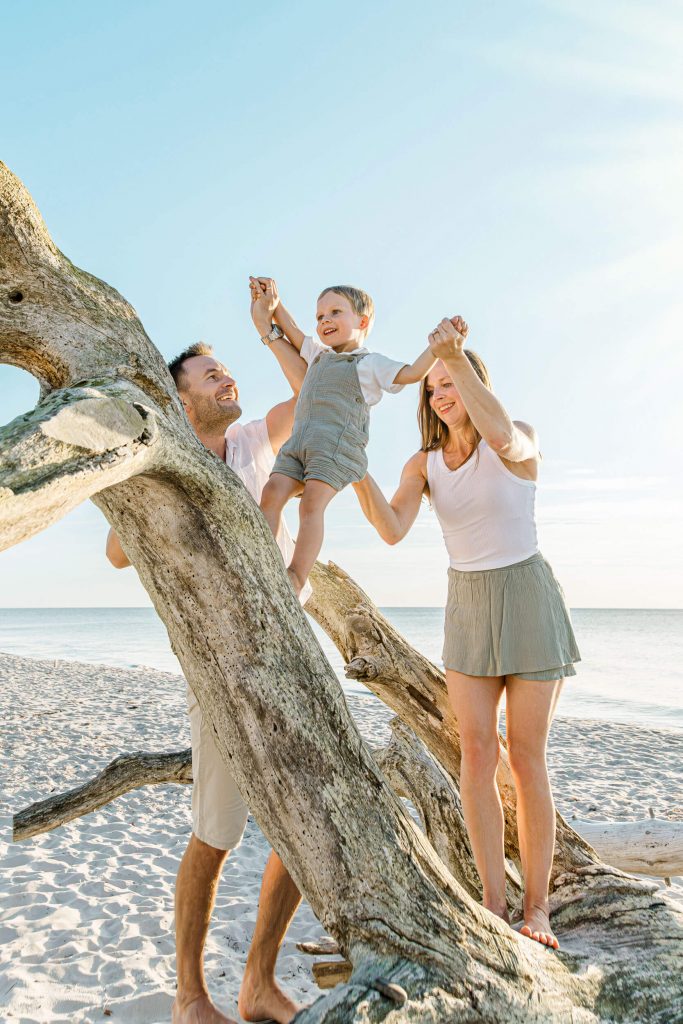 Familie mit einem kleinen Kind, das an einem Baumstamm am Strand spielt, mit klarem Himmel im Hintergrund.