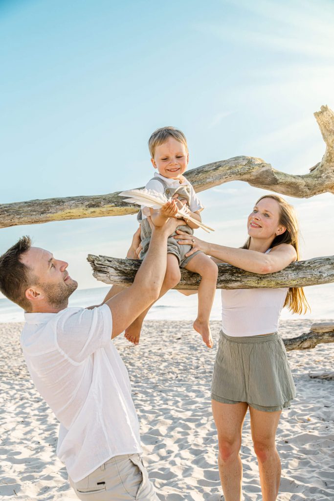 Familie spielt am Strand mit einem Kind, das auf einem Baumstamm sitzt, unter blauem Himmel und Sonnenschein.