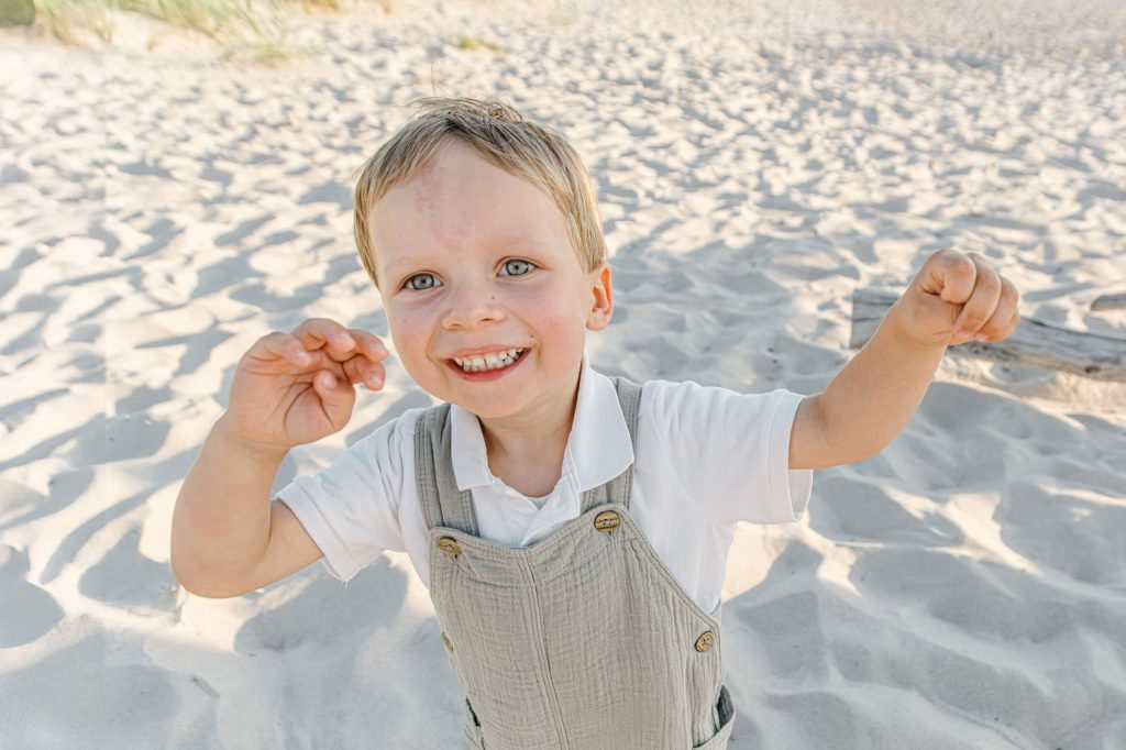 Ein fröhlicher Junge mit blonden Haaren und einem grauen Latzkleid steht auf dem Sandstrand und lächelt in die Kamera.